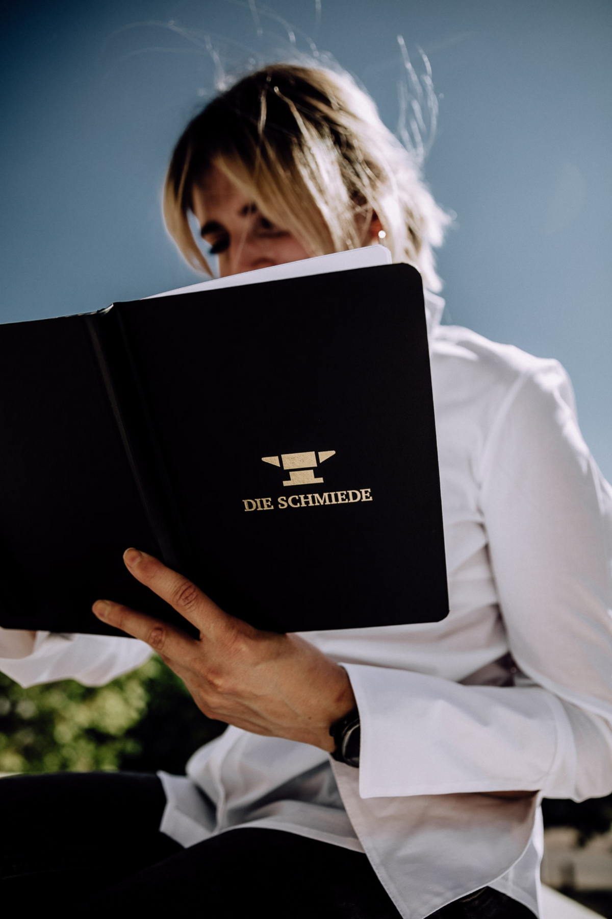 Frau hält schwarzes Notizbuch mit unlinierten Blättern und goldenem DIE SCHMIEDE Logo